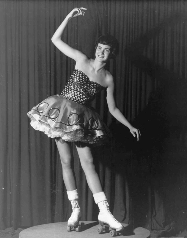 Black-and-white photo of a roller-skating dancer in a patterned short dress, arm raised, smiling against a stage curtain.
